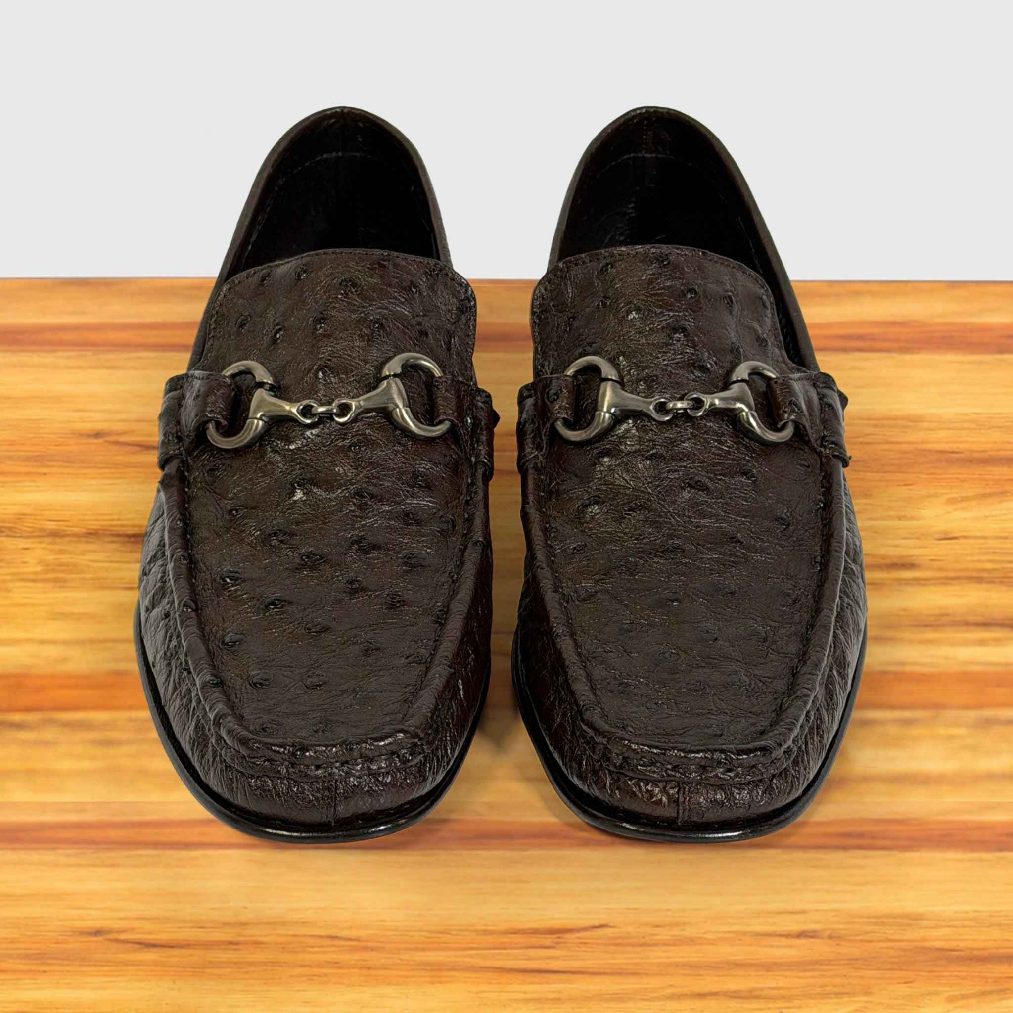 A pair of dark brown ostrich-textured leather loafers , displayed on a wooden surface against a light gray background.