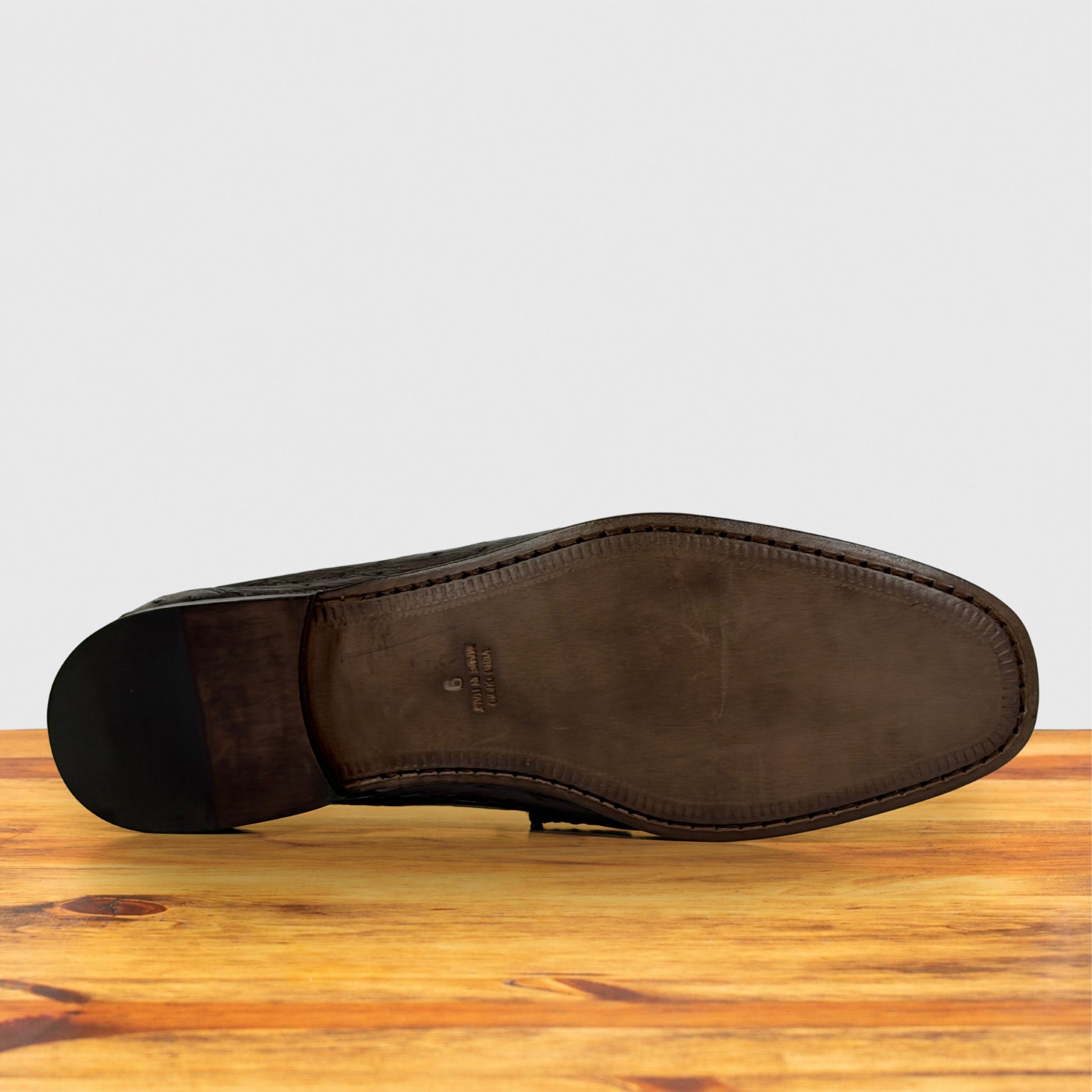 A single dark brown leather shoe shown from the sole side, resting on a wooden surface against a light gray background.