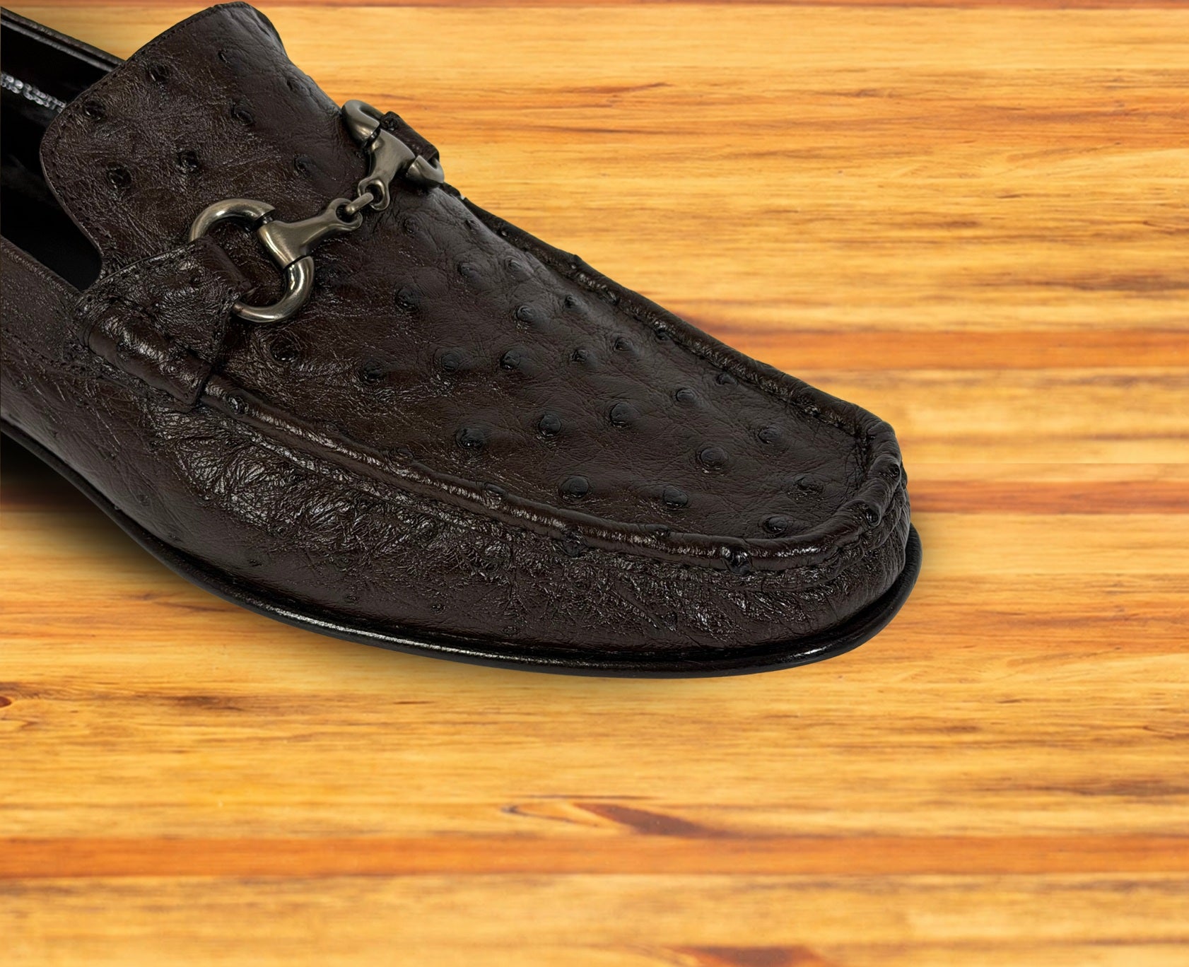 A close-up of a dark brown textured leather loafer with a silver horsebit detail, resting on a light brown wooden surface.