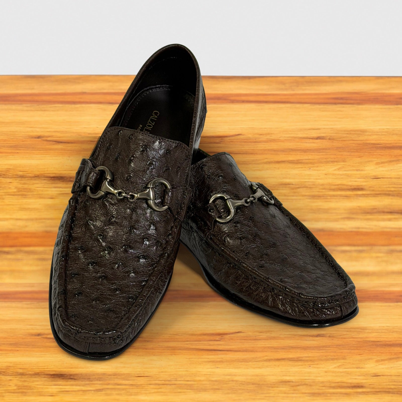 A pair of dark brown textured leather loafers displayed on a wooden surface against a white background.