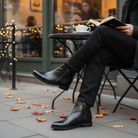 Person wearing black leather boots sitting at an outdoor cafe with a book and coffee cup.