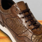 A close-up of a moor color leather sneaker with textured crocodile-embossed panels, dark brown laces, and a white midsole, shown on a wooden surface against a gray background.