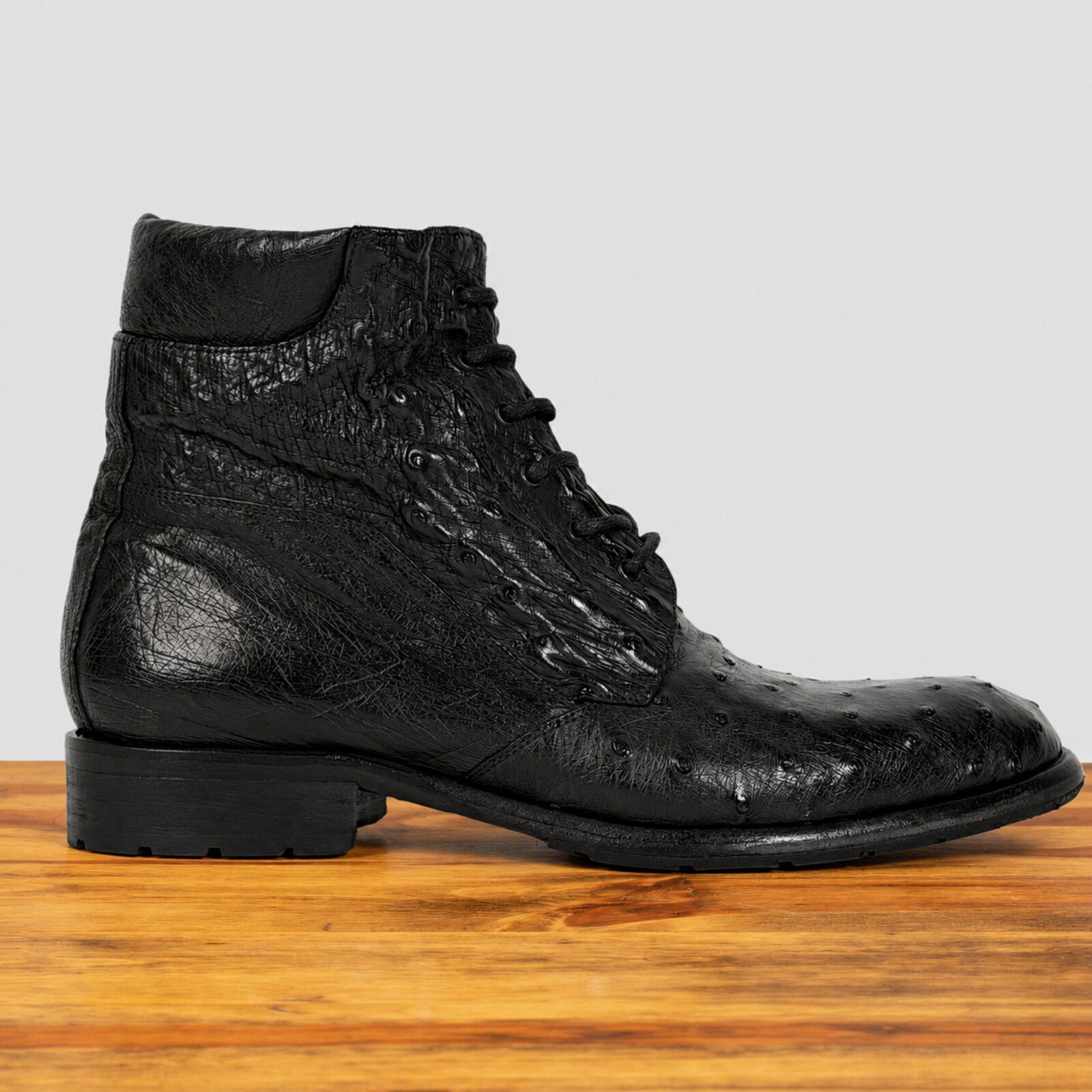 A single black textured lace-up ankle boot with a low block heel, shown in profile on a wooden surface against a light gray background.
