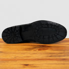 The black rubber outsole of a shoe shown from the bottom, resting on a wooden surface against a light gray background.