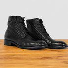 A pair of black textured leather lace-up ankle boots with a glossy finish, displayed on a wooden surface against a light gray background.