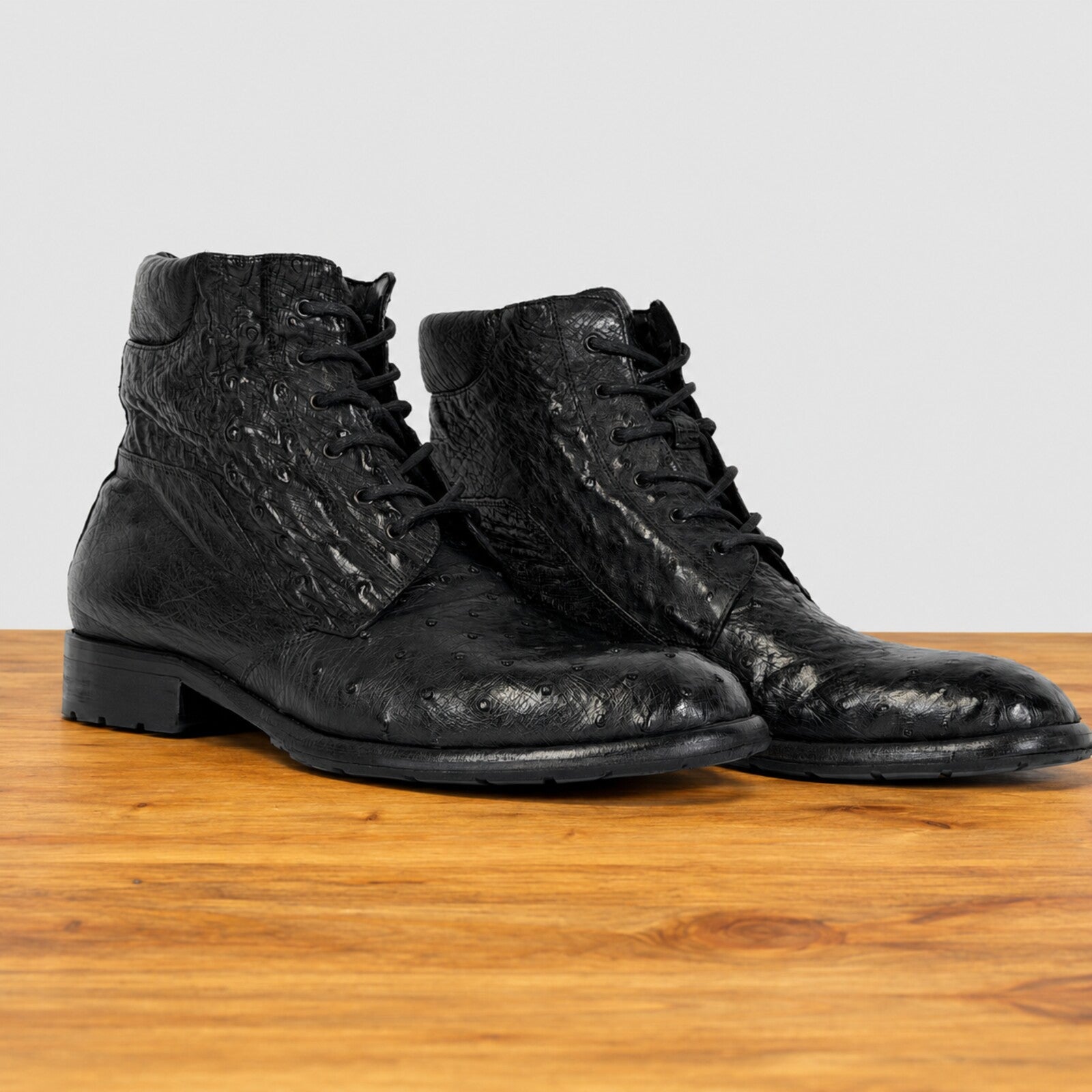A pair of black textured leather lace-up ankle boots with a glossy finish, displayed on a wooden surface against a light gray background.