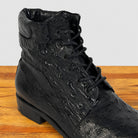 A black textured lace-up ankle boot with an embossed ostrich-style pattern, shown on a wooden surface against a light gray background.