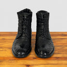 A pair of black textured lace-up ankle boots with a glossy, displayed on a wooden surface against a light gray background.
