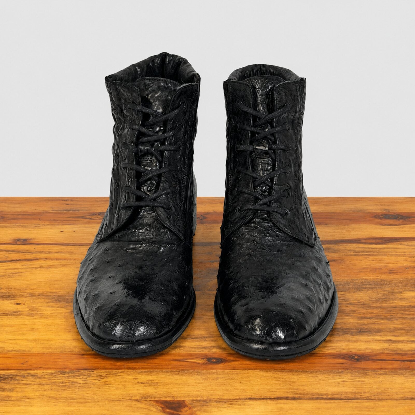A pair of black textured lace-up ankle boots with a glossy, displayed on a wooden surface against a light gray background.