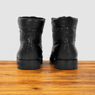 A pair of black leather ankle boots, shown from the back on a wooden surface against a light gray background.