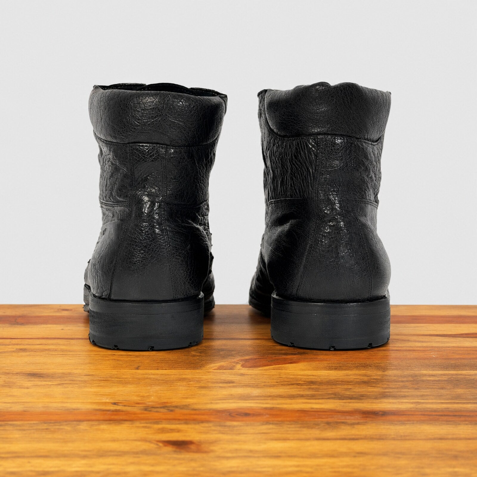 A pair of black leather ankle boots, shown from the back on a wooden surface against a light gray background.