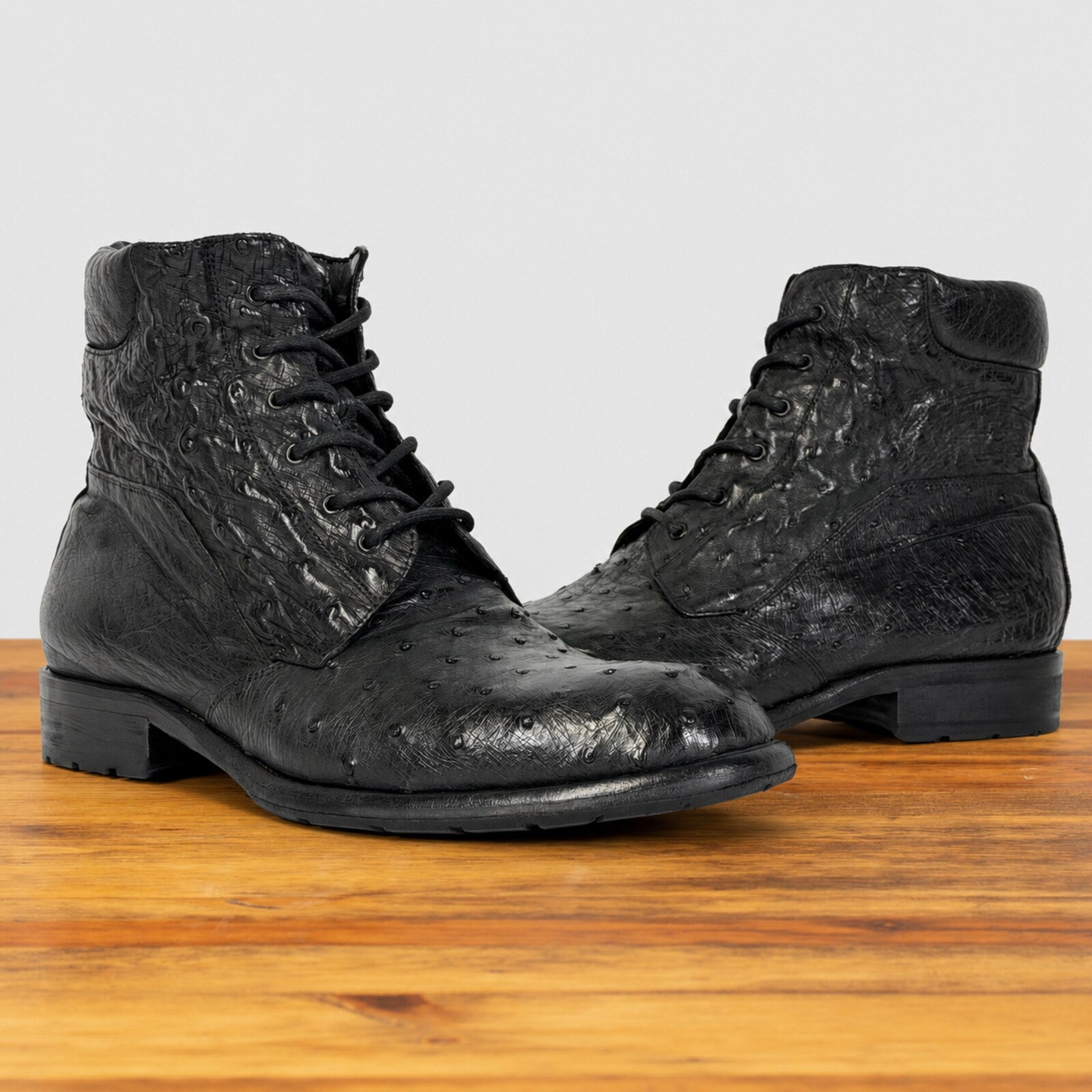 A pair of black lace-up ankle boots with a textured ostrich leather finish, displayed on a wooden surface against a light gray background.