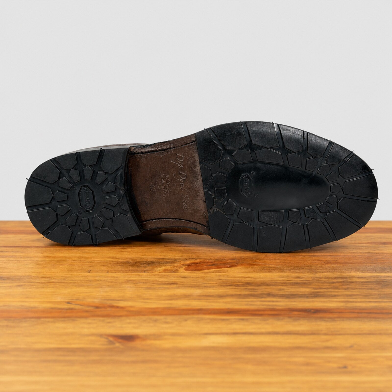 The treaded black rubber outsole of a brown boot is shown from a bottom-up view, resting on a wooden surface against a light gray background.
