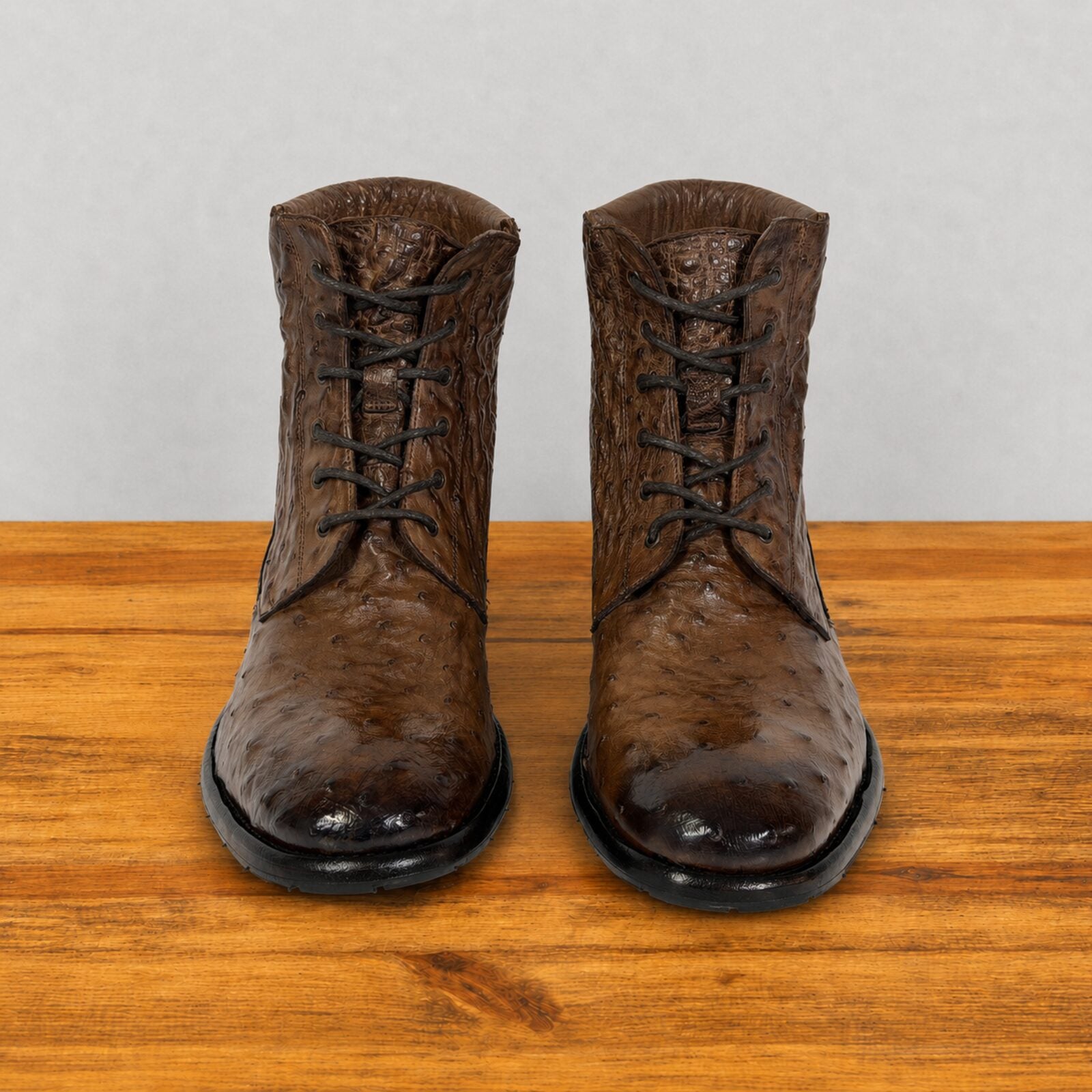 A pair of dark brown textured lace-up boots with black soles, displayed on a wooden surface against a light gray background.