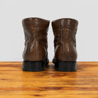 A pair of brown leather ankle boots with black soles, shown from the back on a wooden surface against a light gray background.