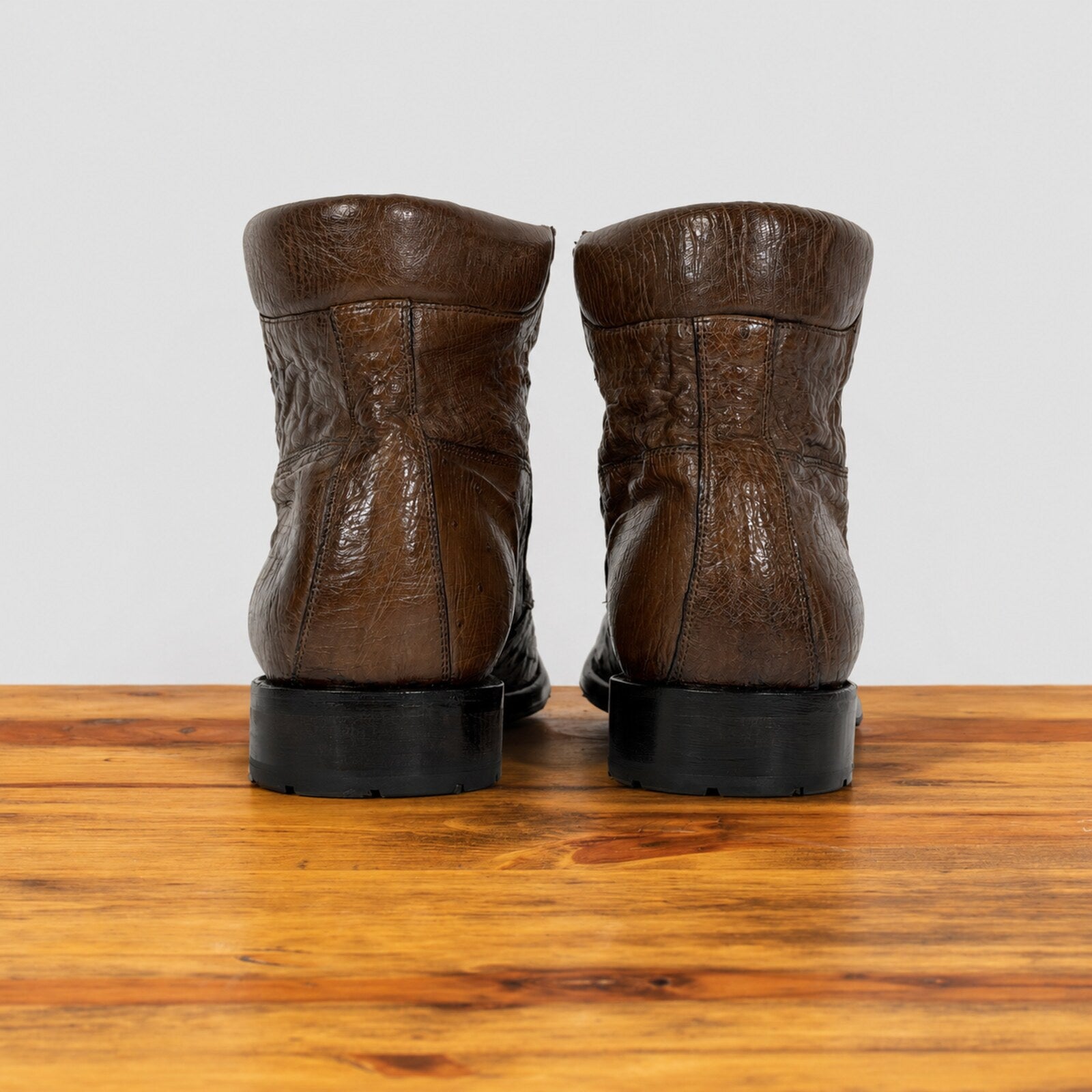 A pair of brown leather ankle boots with black soles, shown from the back on a wooden surface against a light gray background.