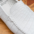 A close-up of a white crocodile-embossed leather loafer on a wooden surface, showing the textured leather, stitching, and part of the white insole with 'DRAFTED SHOES MADE IN ITALY' text.