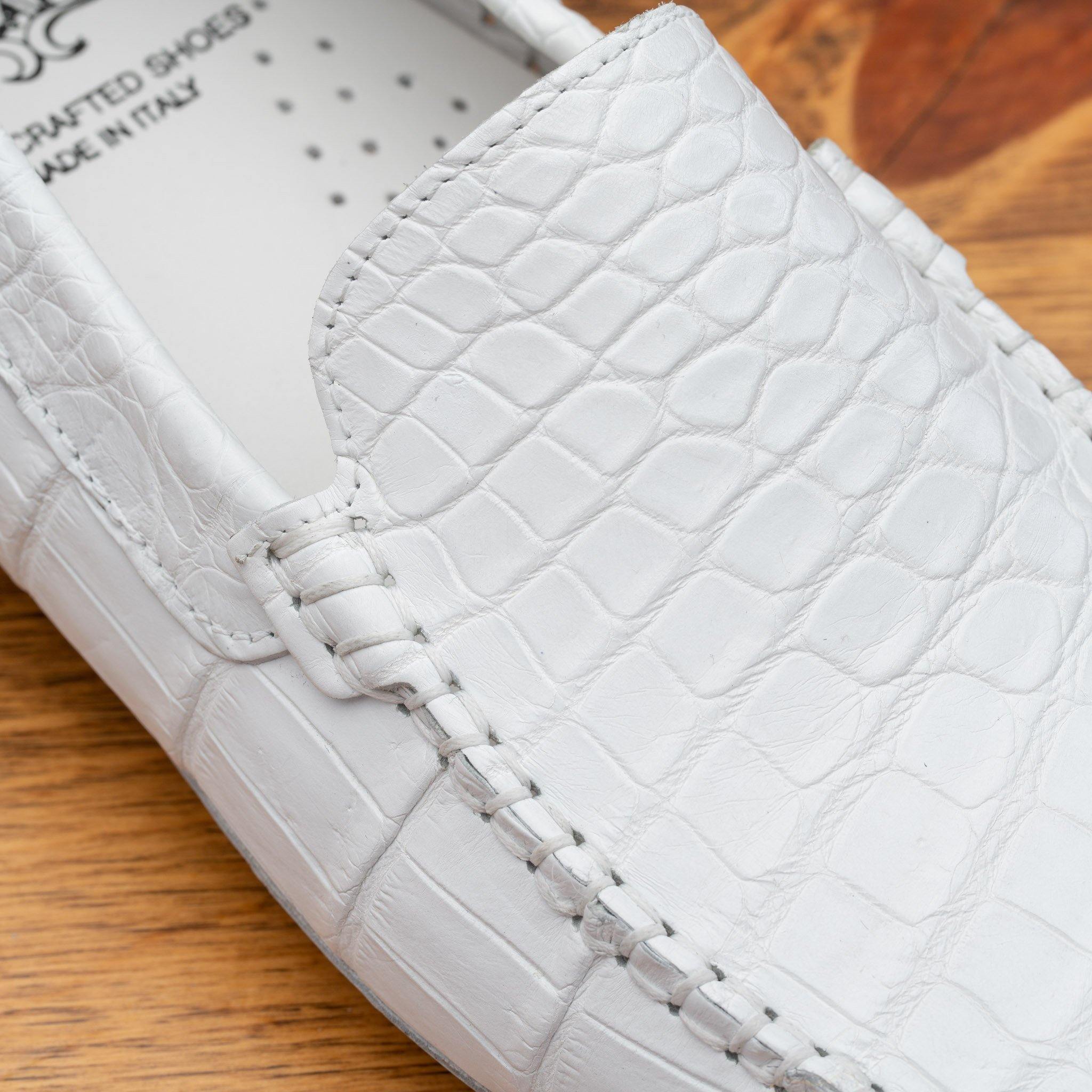 A close-up of a white crocodile-embossed leather loafer on a wooden surface, showing the textured leather, stitching, and part of the white insole with 'DRAFTED SHOES MADE IN ITALY' text.