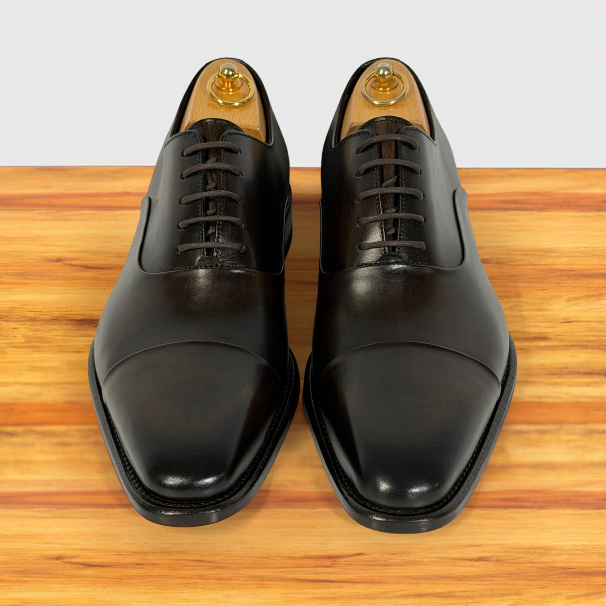 A pair of Dark Brown leather cap-toe oxford shoes with laces, displayed on wooden shoe trees and set on a wooden surface against a light gray background.