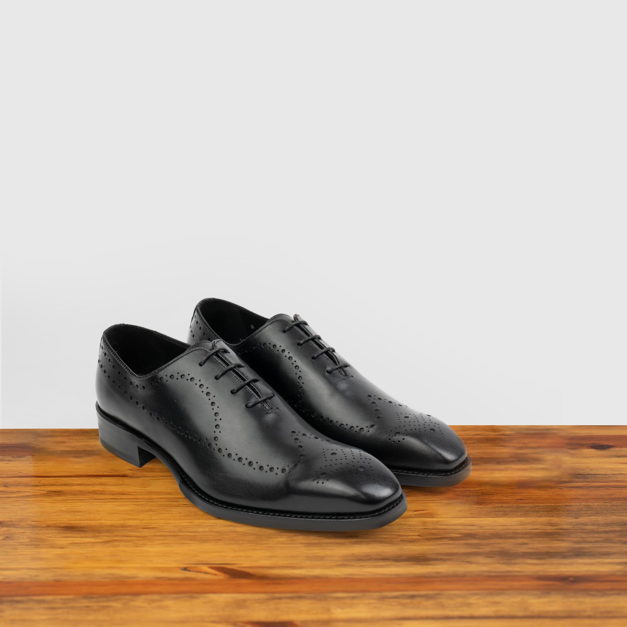 A Pair of Black wholecut dress shoe on a wooden surface with a light gray background