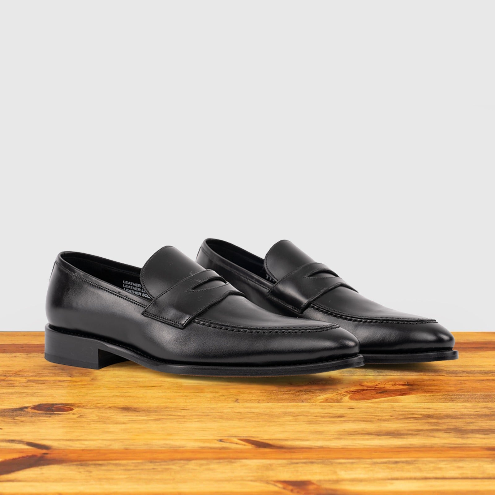 A Pair of black Italian leather dress loafer with a penny strap, side view, on a wooden surface.