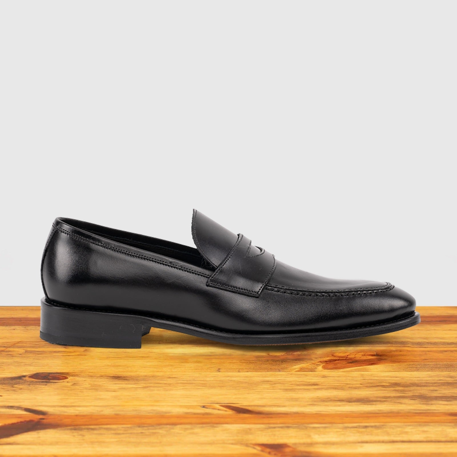 A black Italian leather dress loafer with a penny strap, side view, on a wooden surface.