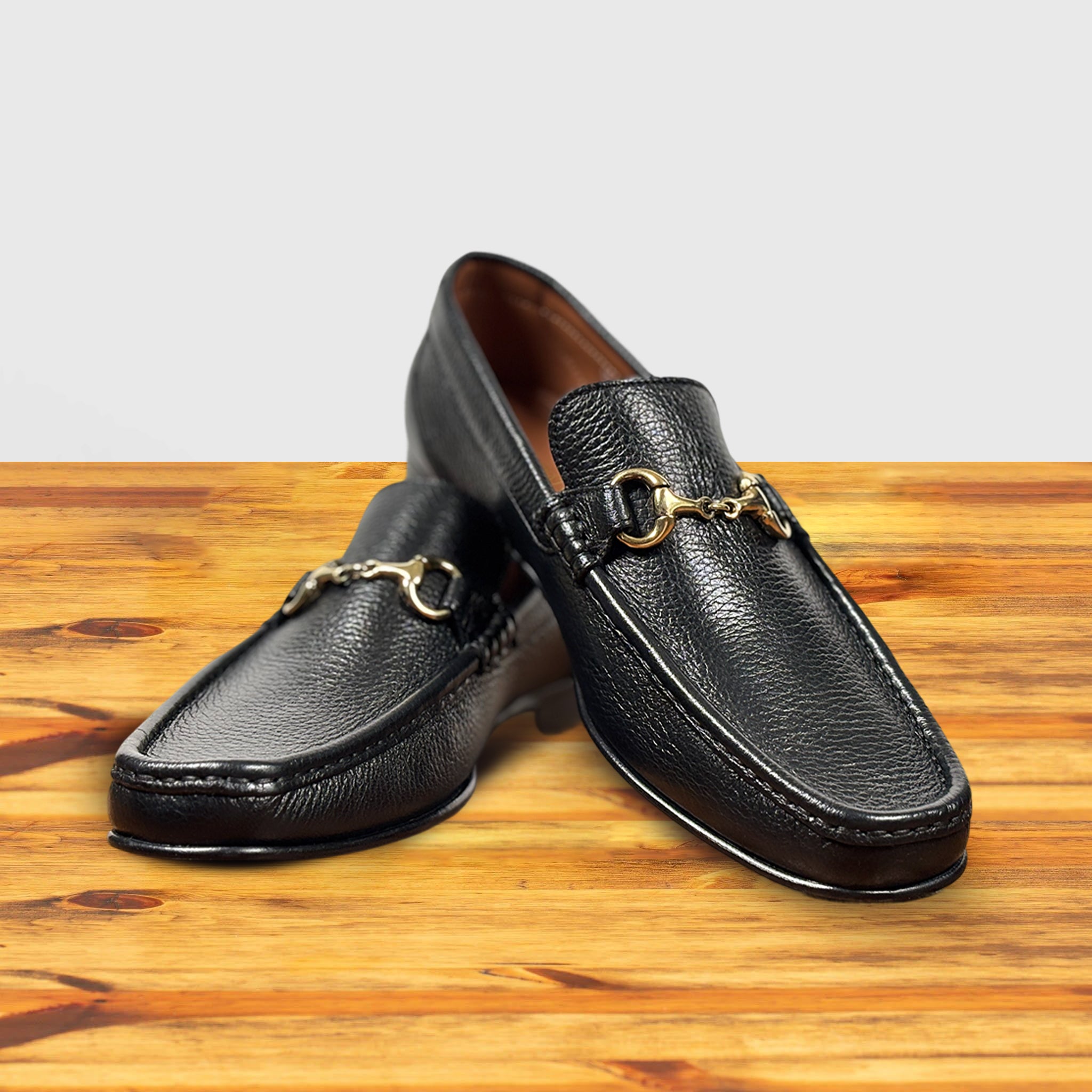 A pair of Black leather loafer with a gold buckle on a wooden surface in a light gray background