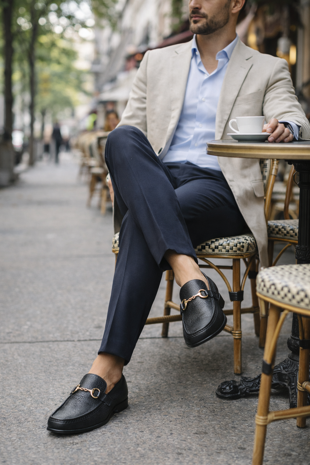 Man sitting at a cafe table wearing a light gray blazer, blue shirt, dark pants, and black loafers.
