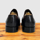 A pair of blue leather wingtip oxford shoes with brogue detailing, shown from the back on a wooden surface against a light gray background.