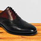 A men's wingtip oxford shoe in blue and dark brown leather, featuring decorative brogue perforations, a black lace, and a black sole, displayed on a wooden surface against a light gray background.