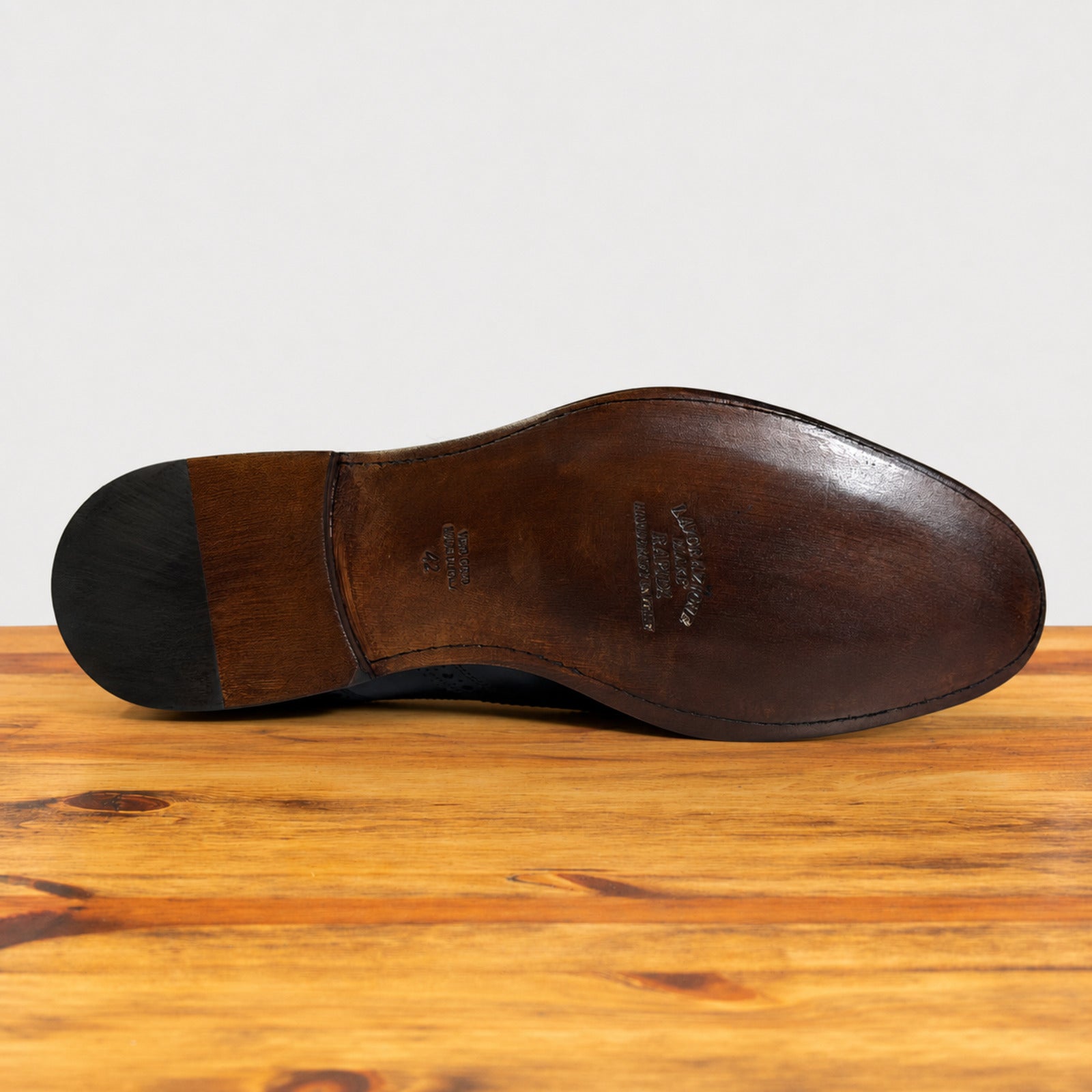 The outsole of a leather dress shoe, showing the brand name 'BOSTON SHOES' and size markings, resting on a wooden surface against a light gray background.