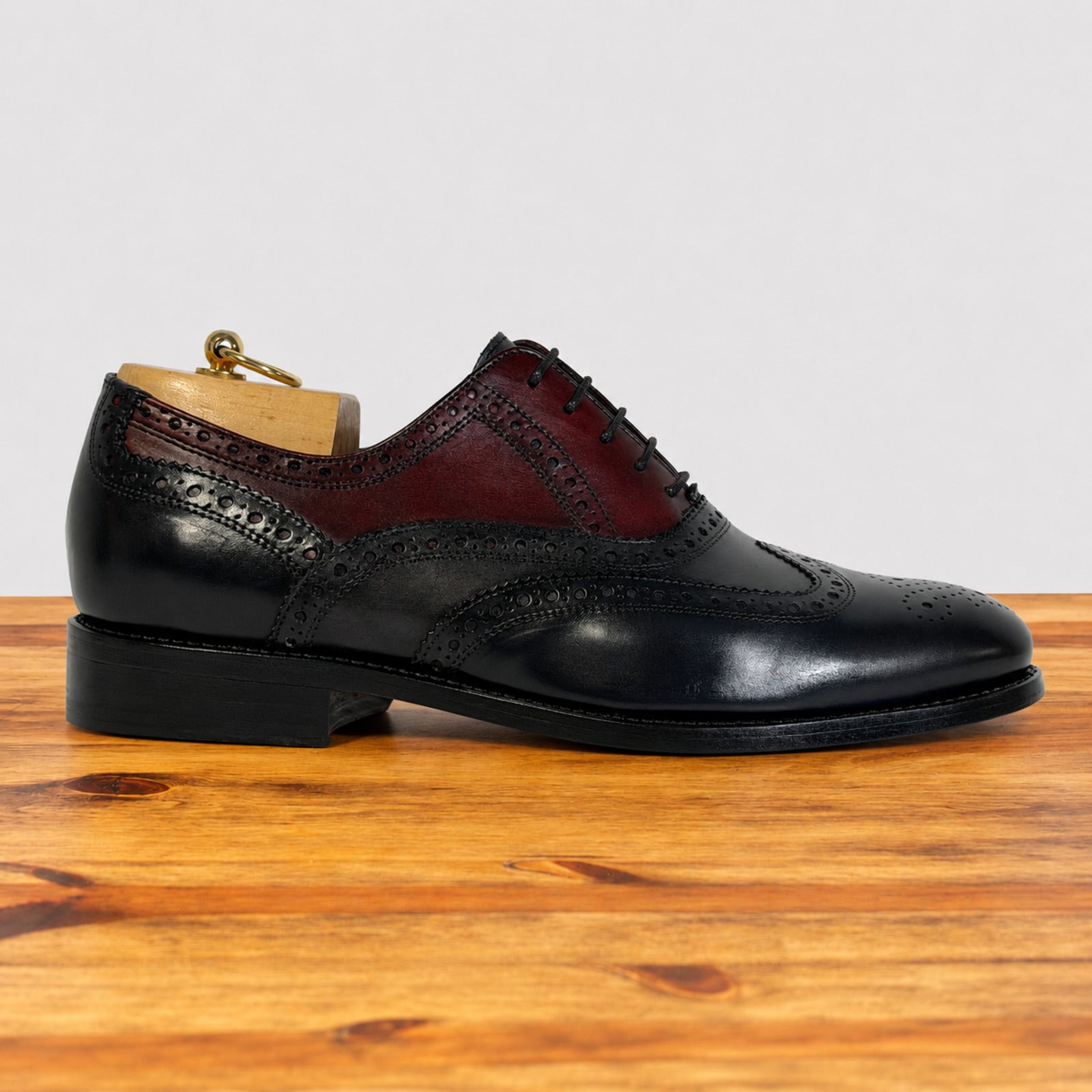 A men's wingtip oxford shoe in midnight blue and gray leather, displayed on a wooden surface against a light gray background. The shoe is fitted with a wooden shoe tree.