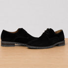 A pair of men's black suede derby shoes with dark speckled soles, displayed on a light wood surface against a neutral light gray background.