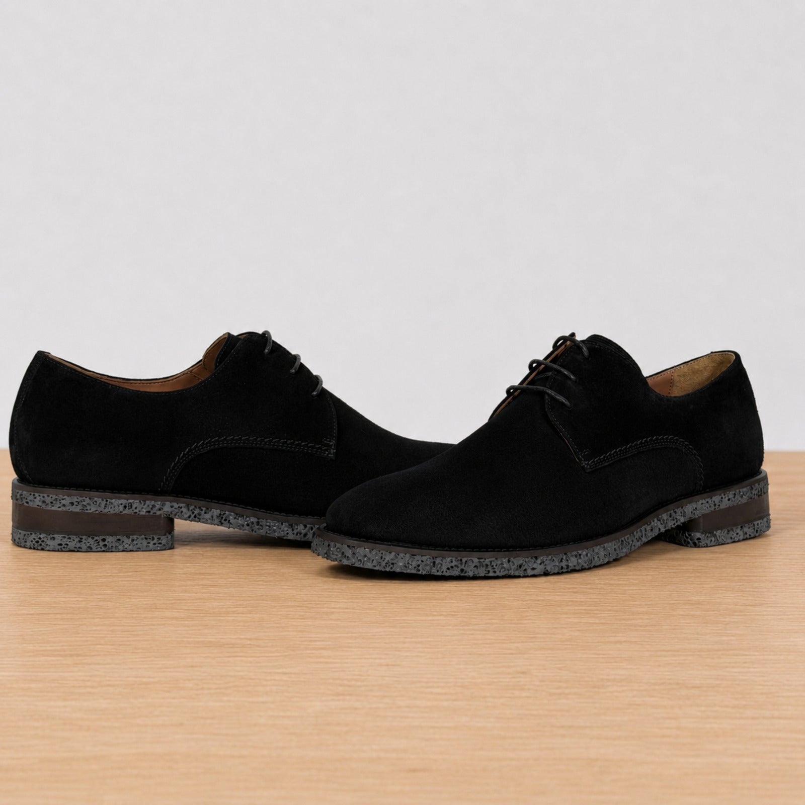 A pair of men's black suede derby shoes with dark speckled soles, displayed on a light wood surface against a neutral light gray background.