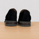 A pair of black suede shoes shown from the back, resting on a light wood surface against a plain light gray background.