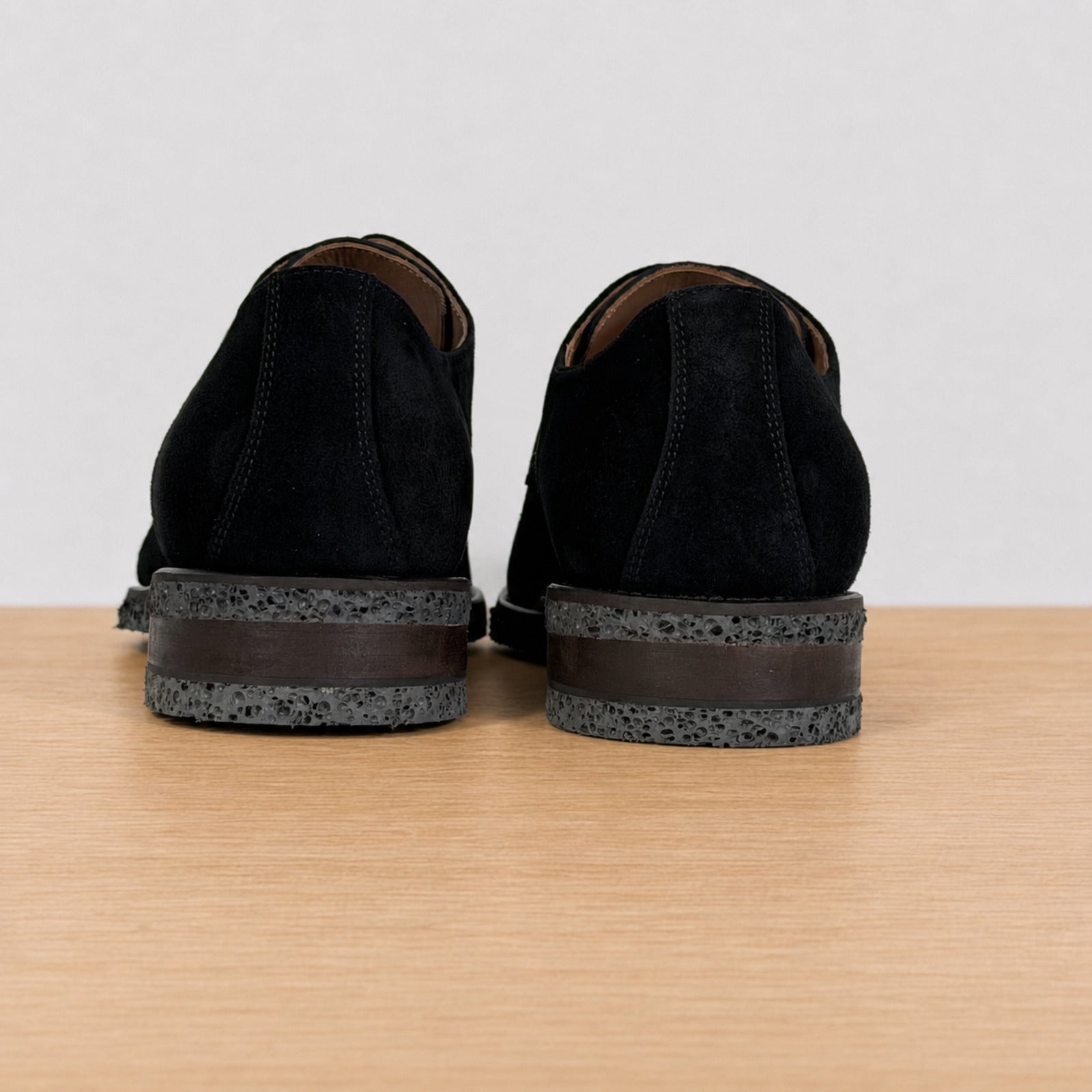 A pair of black suede shoes shown from the back, resting on a light wood surface against a plain light gray background.