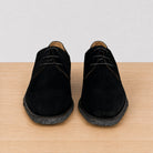 A pair of black suede lace-up shoes with textured black soles, displayed from a front-facing perspective on a light wood surface against a white background.