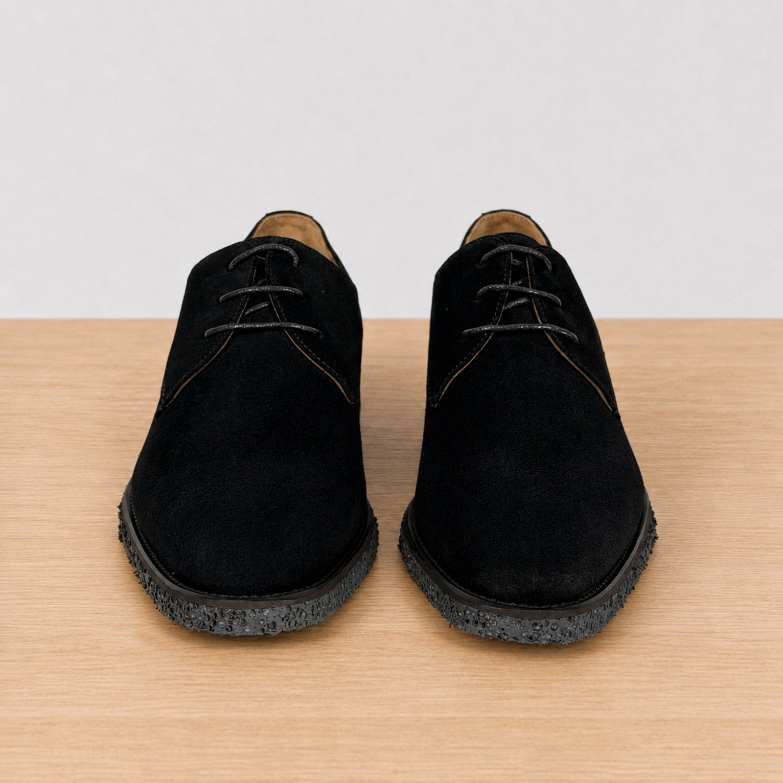 A pair of black suede lace-up shoes with textured black soles, displayed from a front-facing perspective on a light wood surface against a white background.