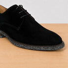 A black suede derby shoe with black laces and a speckled dark gray sole, shown on a light wood surface against a white background.