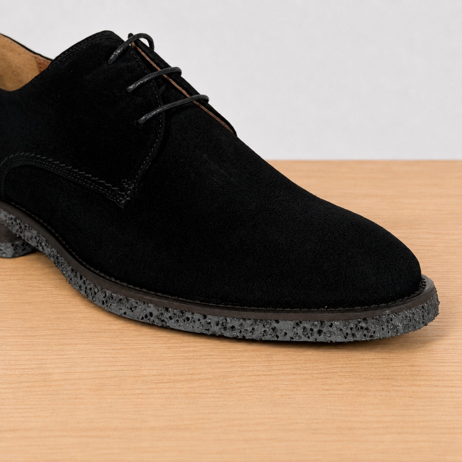 A black suede derby shoe with black laces and a speckled dark gray sole, shown on a light wood surface against a white background.