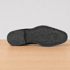 The textured black outsole of a shoe resting on a light wood surface against a neutral light gray background.