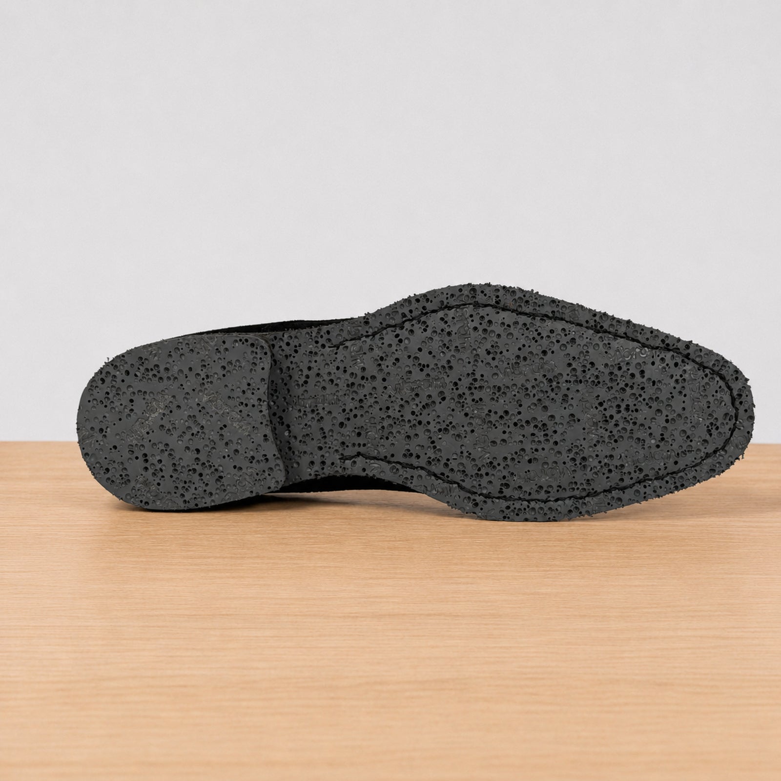 The textured black outsole of a shoe resting on a light wood surface against a neutral light gray background.