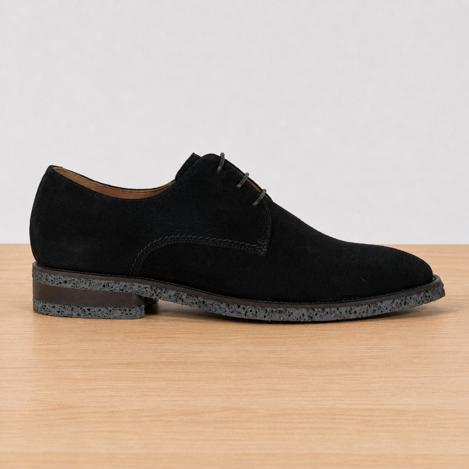 A single black suede derby shoe with a speckled dark sole, shown in profile on a light wood surface against a white background.