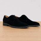 A pair of black suede derby shoes with black laces and speckled black soles, displayed on a light wood surface against a neutral light gray background.