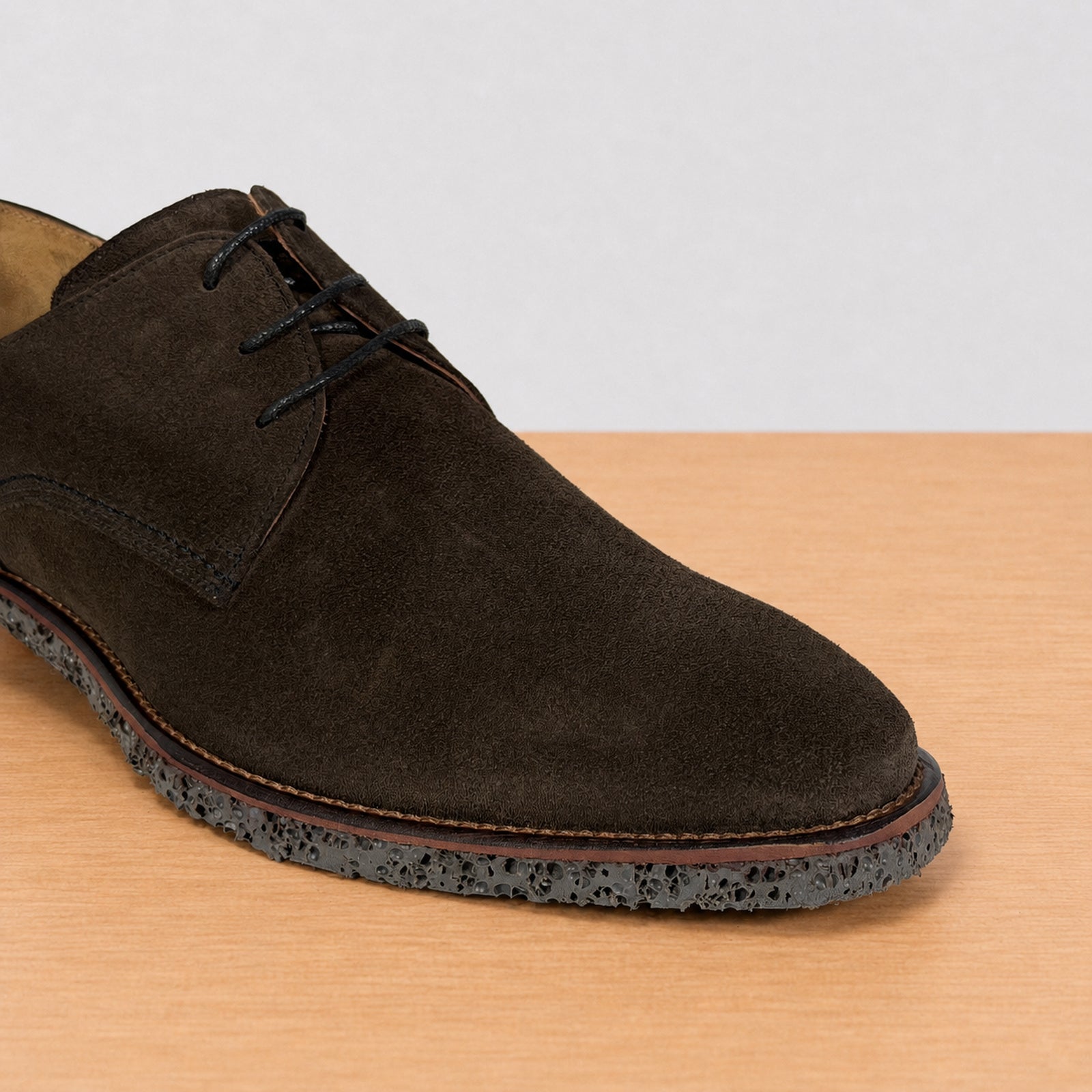 A dark brown suede lace-up shoe with a speckled black rubber sole, shown on a light wood surface against a neutral light gray background.
