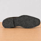 The textured black outsole of a shoe shown from a side profile view, resting on a light wood surface against a neutral light gray background.