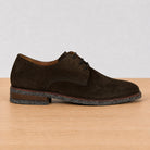 A dark brown suede derby shoe with black laces, shown in a side profile on a light wood surface against a neutral light gray background.