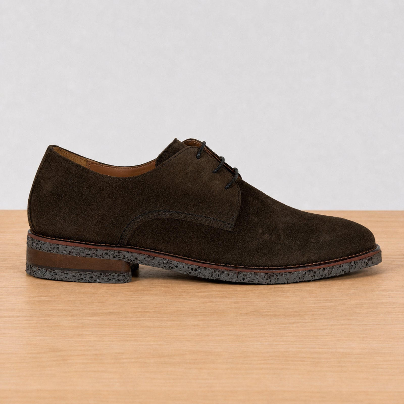 A dark brown suede derby shoe with black laces, shown in a side profile on a light wood surface against a neutral light gray background.