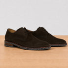 A pair of dark brown suede derby shoes with laces and speckled soles, displayed on a light wood surface against a neutral light gray background.
