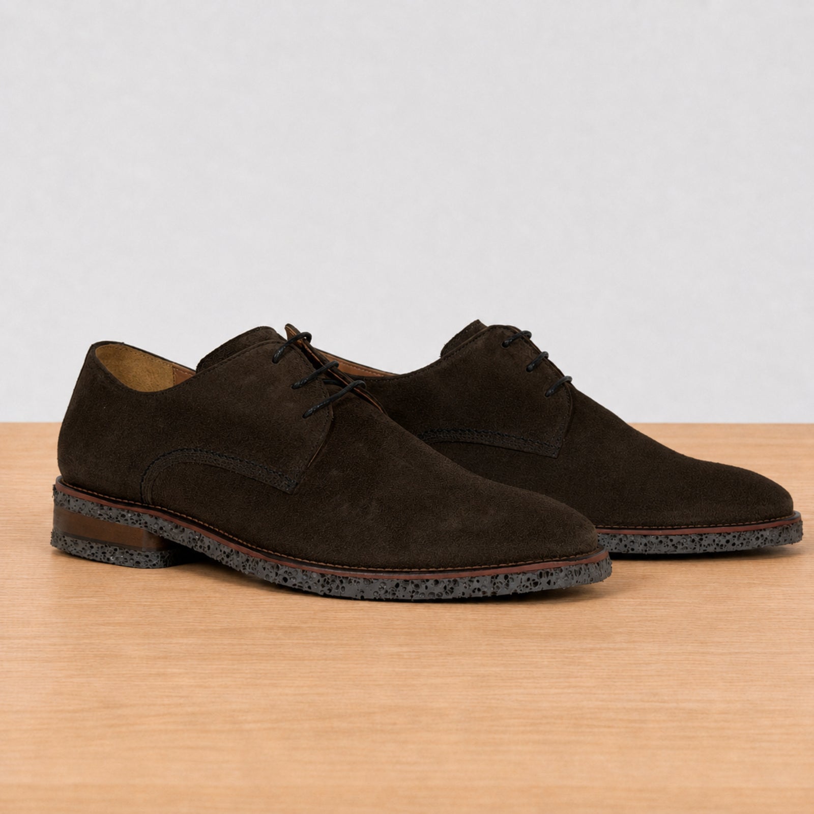 A pair of dark brown suede derby shoes with laces and speckled soles, displayed on a light wood surface against a neutral light gray background.